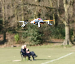 SQ1 mini quadcoper flying against spring treetops with an elderly couple on a park bench watching from below